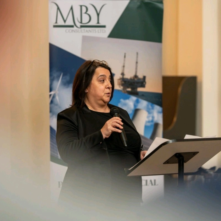 A woman speaks at a podium in front of a backdrop featuring an oil rig.