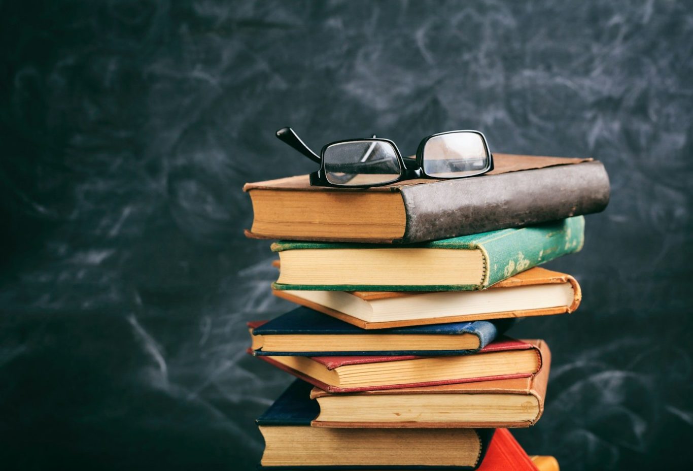 Stack of books with a pair of glasses resting on top, against a chalkboard background.