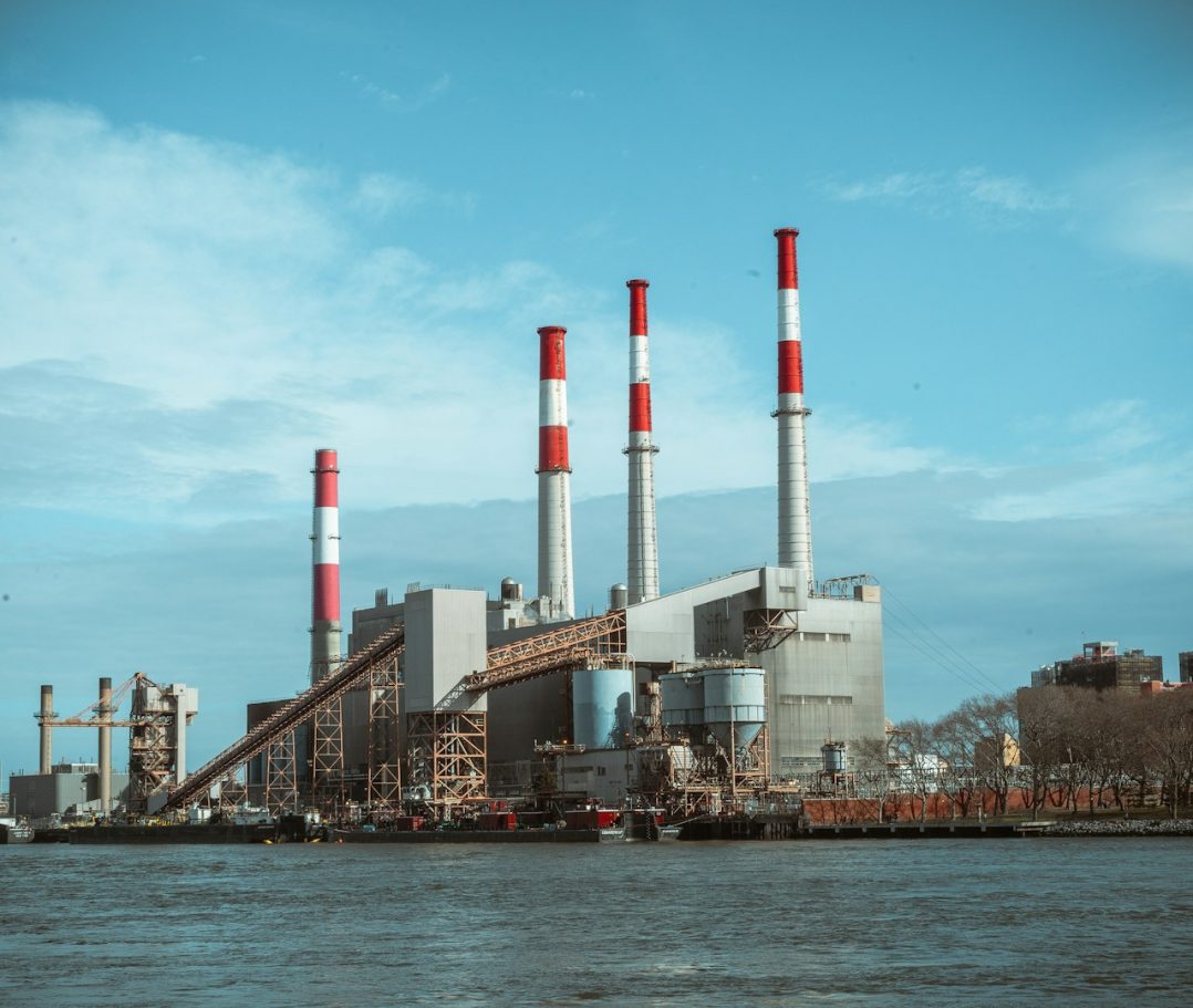 Industrial power plant with tall red and white smokestacks by a river under a blue sky.