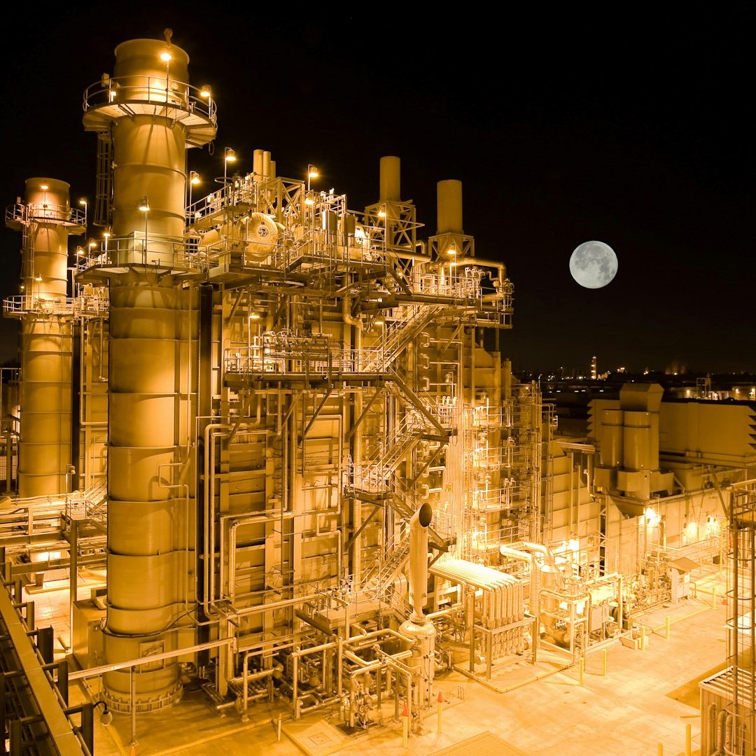 Nighttime industrial plant with illuminated machinery and a large moon in the sky.